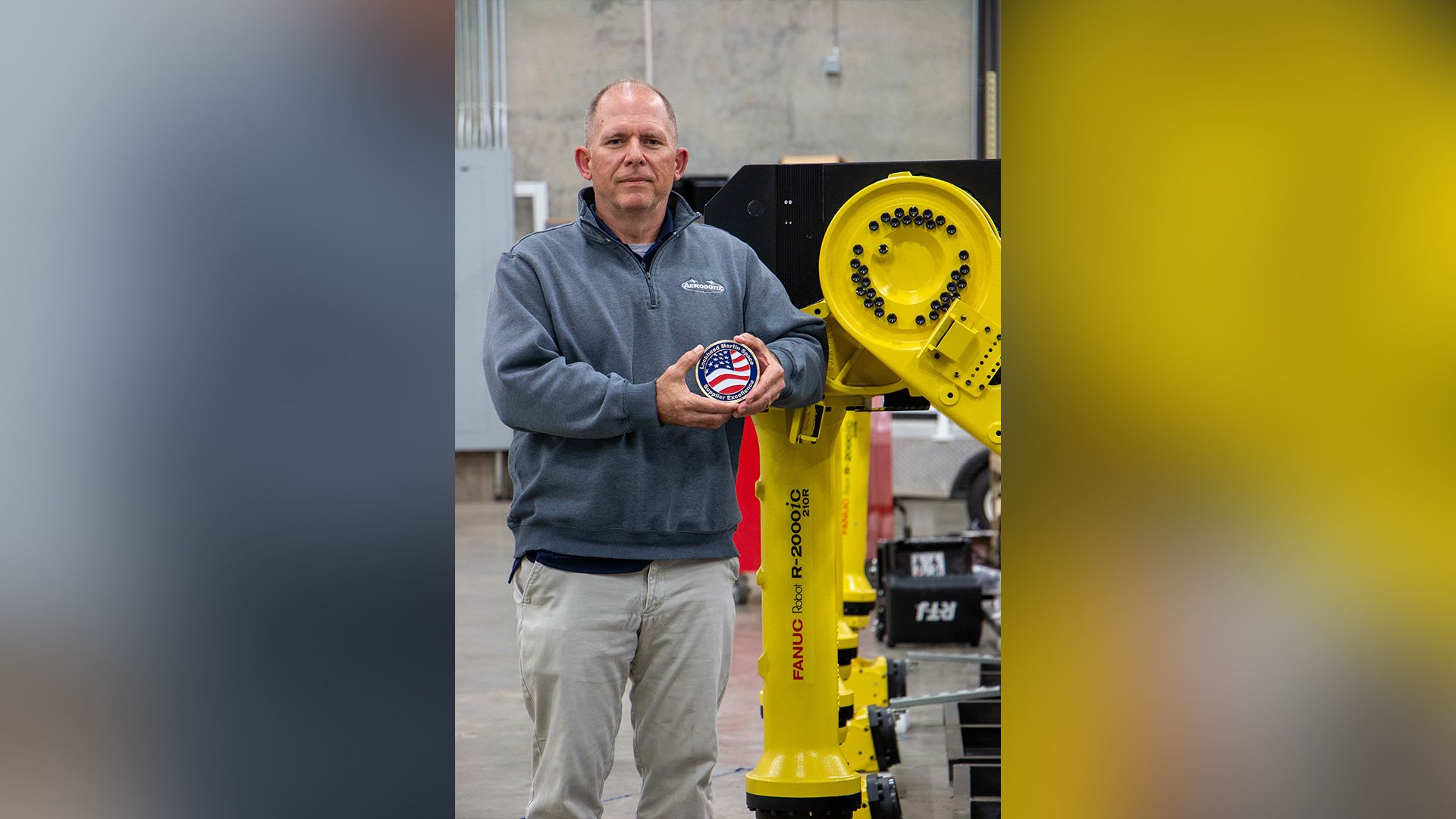 Aerobotix Senior Project Engineer Keenan Simmons holding the 2022 Lockheed Martin Supplier Recognition Award for Ease of Doing Business beside a FANUC robot.